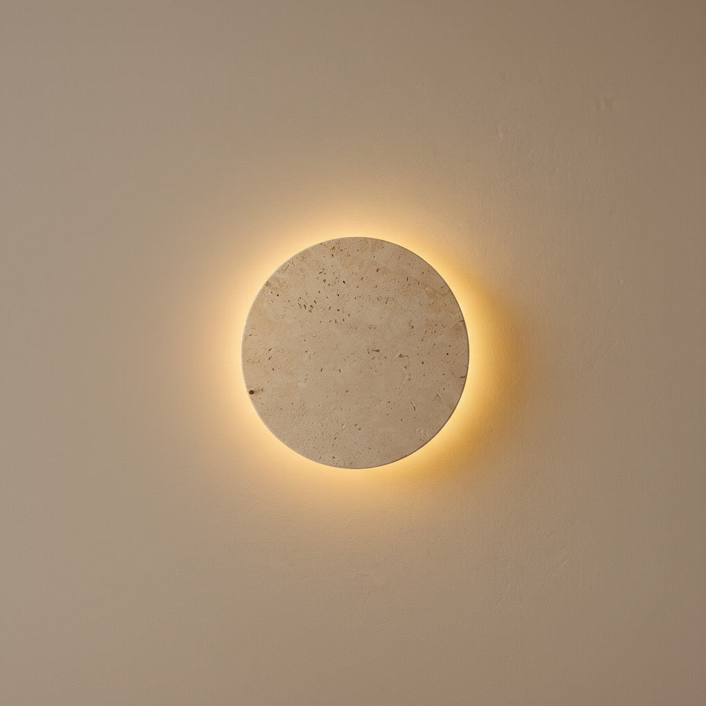 Applique disque Ishi en travertin allumée, halo de lumière sur un mur clair, fond neutre